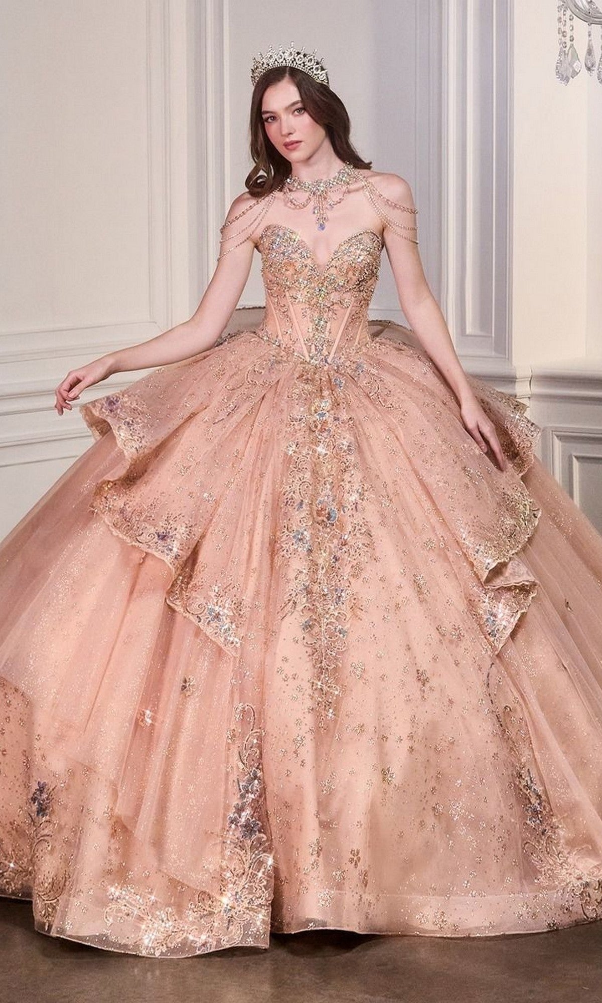 A woman stands in an elegant room wearing the Quinceanera Dress 15727 by Ladivine, a rose gold ball gown with jeweled details and intricate beaded embroidery.